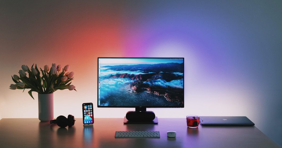 Modern desk setup with monitor displaying mountain landscape, smartphone, flowers, and keyboard